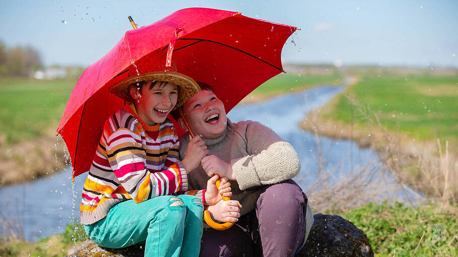 Zwei Freunde lachen unter einem Regenschirm im Regen. Kinder werden im Freien vom Regen überrascht.