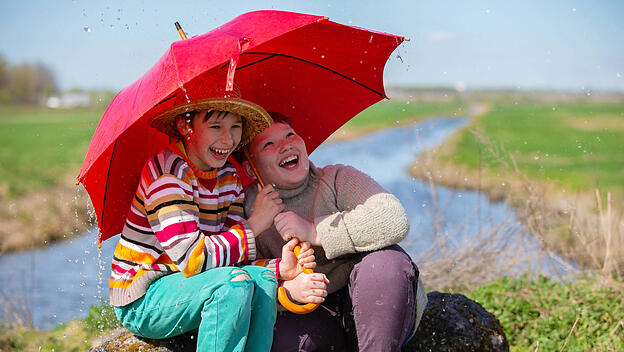 Zwei Freunde lachen unter einem Regenschirm im Regen. Kinder werden im Freien vom Regen überrascht.