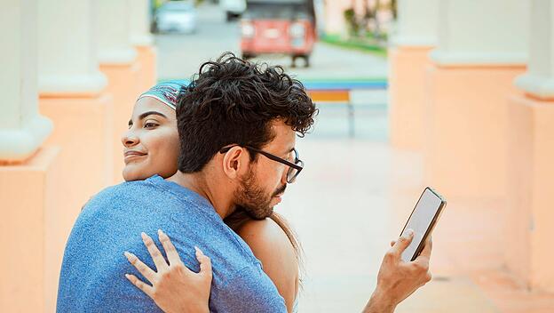 Mann schaut auf Handy, w&auml;hrend er seine Frau umarmt