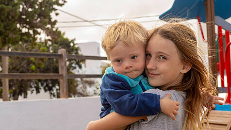 Young girl embracing her toddler brother with Down syndrome at a playground, showing affection and inclusion. Model Rele
