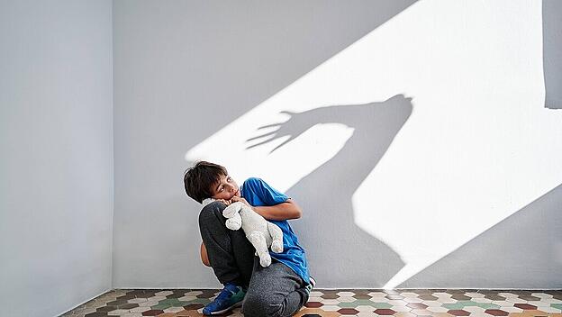 Scared little boy with toy in hands sitting near wall with shadow of angry violent parent with raised arm, Model release