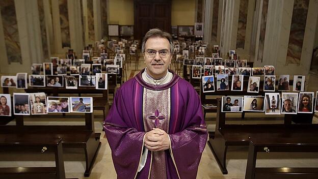 Pfarrer Don Giuseppe Corbari steht vor Kirchenbänken mit ausgedruckten Fotos der Kirchengemeinde