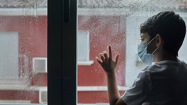 Unhappy child wearing face mask standing near window and observing street while staying home during coronavirus epidemic