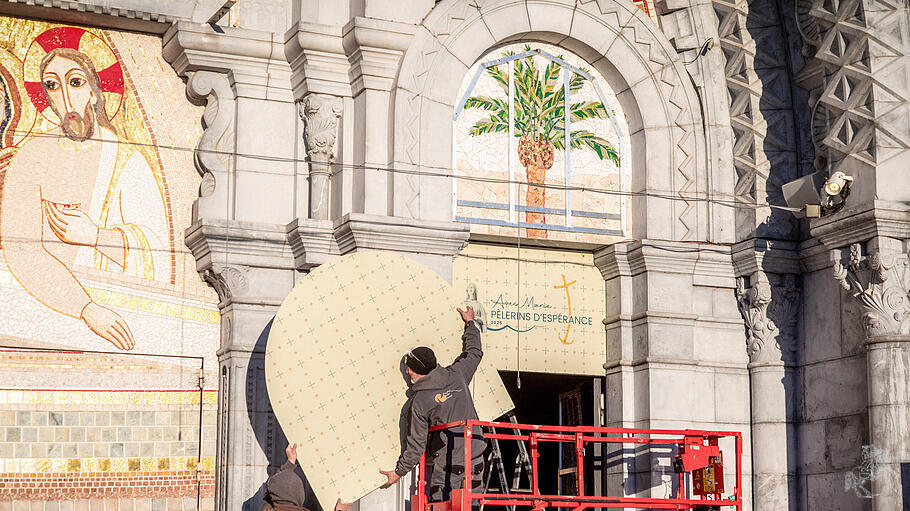 Arbeiter halten eine Abdeckung, die zur Verhüllung des Mosaiks am Eingang der Rosenkranz-Basilika angebracht wird, am 31. März 2025 in Lourdes (Frankreich). Das Mosaik ist ein Kunstwerk von Priester und Künstler Marko Ivan Rupnik, der unter Verdacht steht, sexuellen Missbrauch begangen zu haben.
